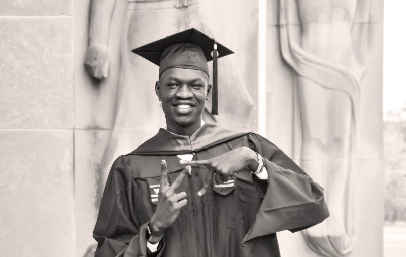 Dudi Miabok, wearing a cap and gown, makes the VT symbol with his hands in front of the Pylons